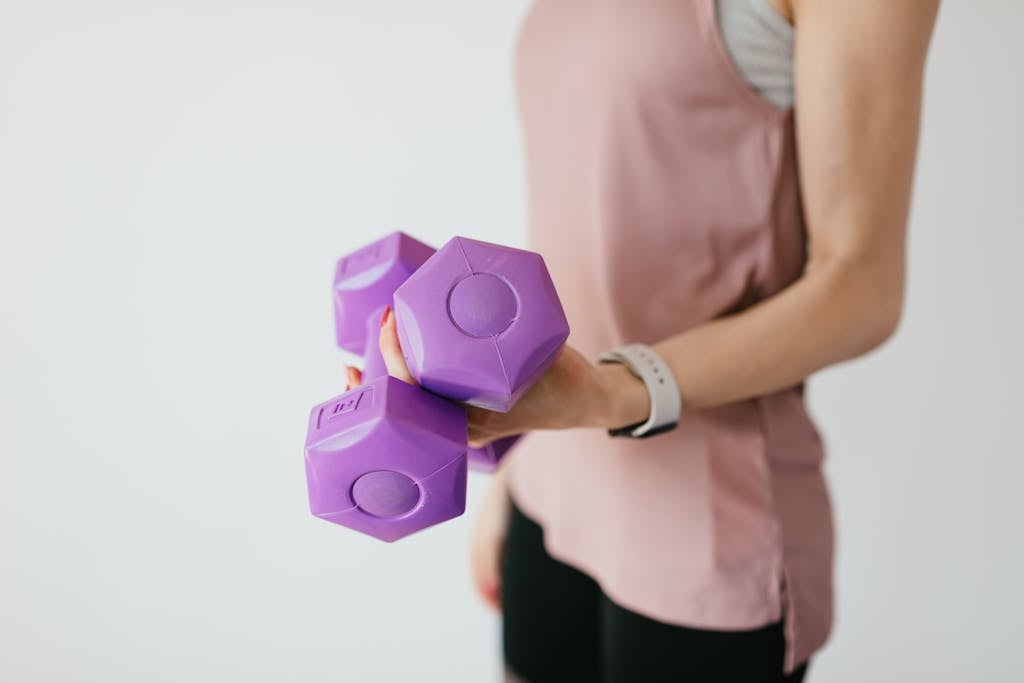 Side view crop anonymous slim female in light pink top and black leggings with lightweight dumbbells against gray wall